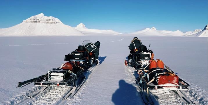 Glasiologisk feltarbeid ved Kongsfjorden. To forskere på hver sin snøskuter med sleder fulle av vitenskapelig utstyr, på vei over isen i solfylt snødekket landskap med fjell i bakgrunnen. Foto: Dagmara Wojtanowicz / NP