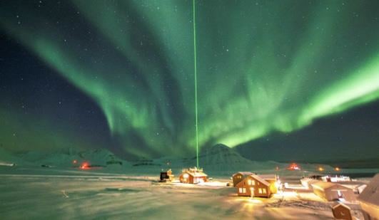 Studier av nordlys i Ny-Ålesund ved bruk av laser. En klynge hus i mørket i snødekket landskap med fjell i bakgrunnen med grønt nordlys og stjerner på himmelen og en grønn laserstråle som går rett opp fra det ene huset i himmelen. Foto: Christelle Guesnon | NP