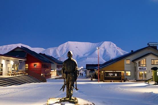 Longyearbyen. En statue av en gruvearbeider står i mørket på et torg med lave trehus med butikker og kafeer på begge sider. Snødekte fjell lyser helt hvitt i bakgrunnen. Foto: Ingrid Ballari Nilssen / UNIS