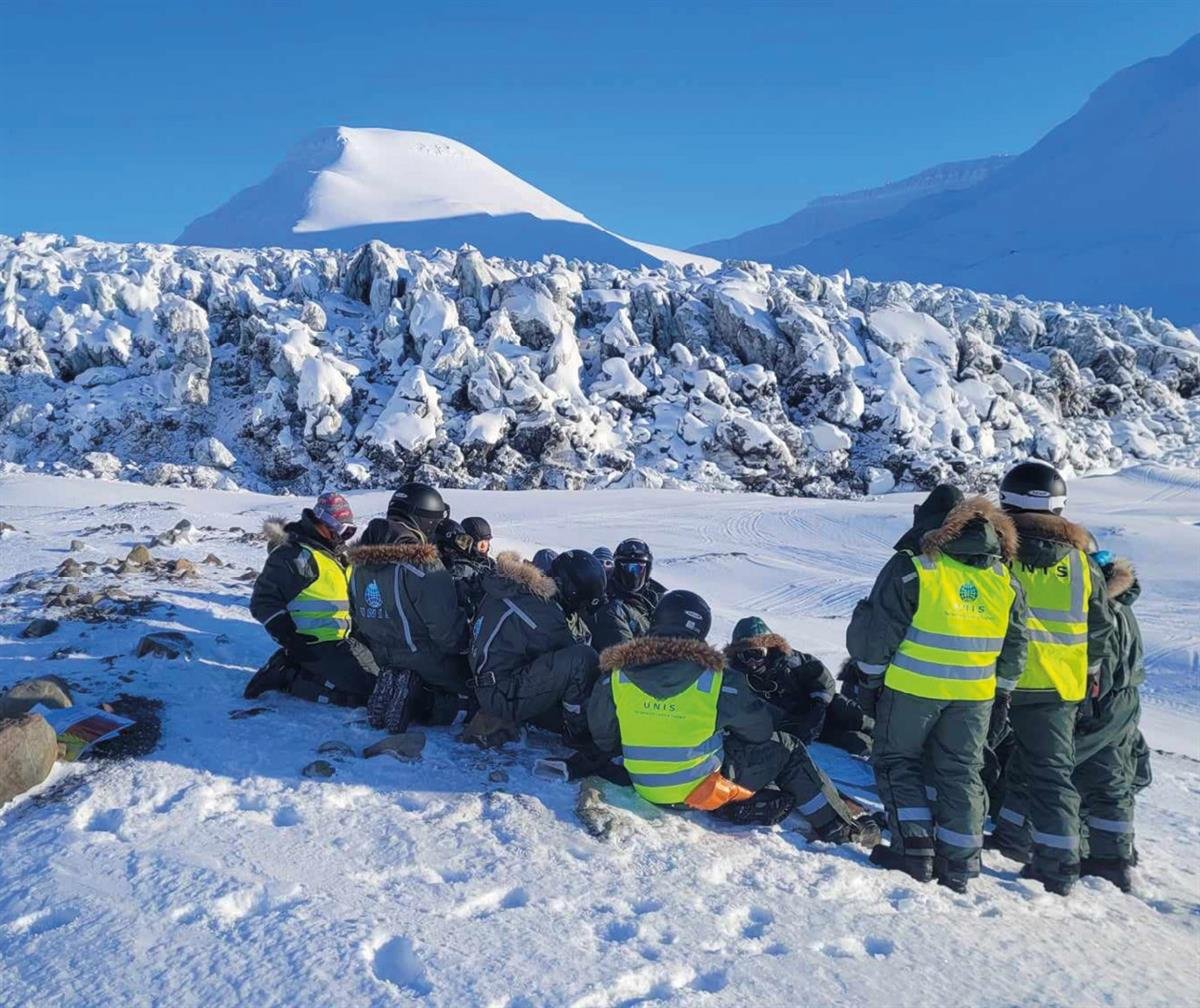En gruppe studenter på feltekskursjon ved Vallåkrabreen i Rindersbukta. Studentene sitter og står i en klynge i et snødekket landskap med Vallåkrabreen og et fjell i bakgrunnen. Foto: Nil Rodes / UNIS