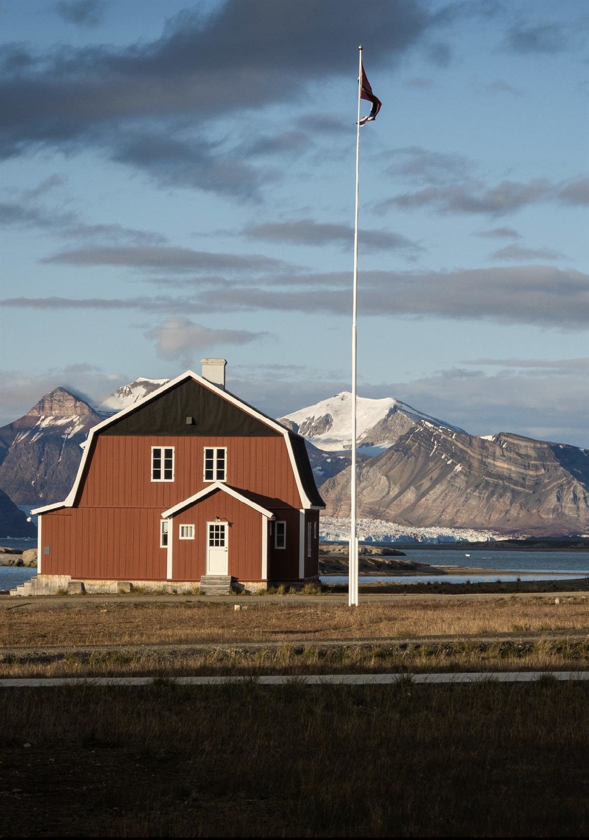 Amundsenvillaen i Ny-Ålesund. Rød trevilla på en gresslette med flaggstang med norsk vimpel, med fjord, isbre og snødekte fjelltopper bak. Fjellene viser de typiske lagdelte bergartene på Svalbard. (Utsnitt) Foto: Trine Lise Sviggum Helgerud / NP