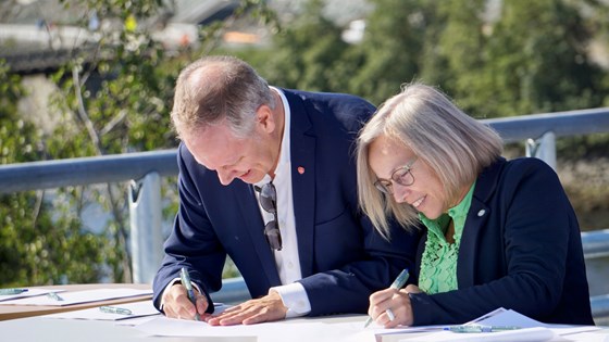 Samferdselsministeren og statssekretæren i KDD står bøyd over et bord for å signere byvekstavtalen for Trondheimsområdet