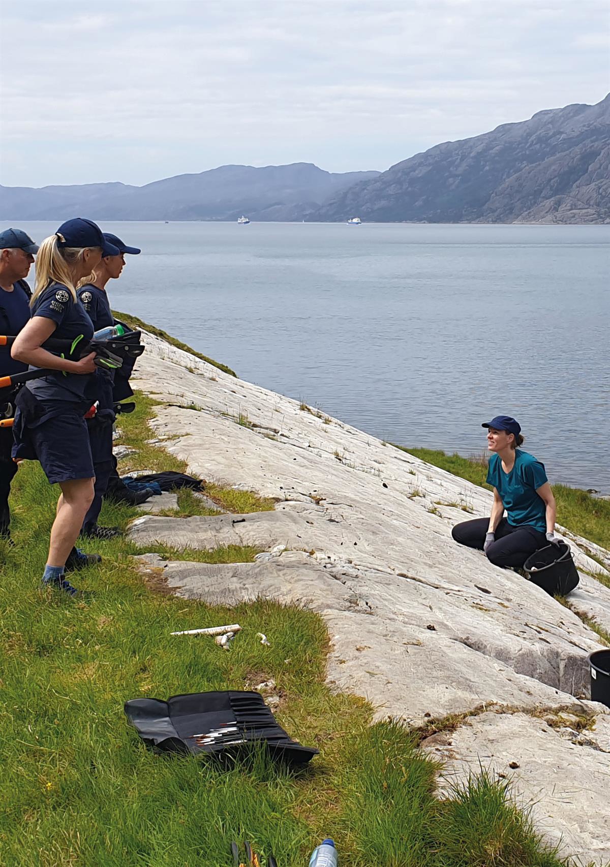 Fire personer i arbeidsklær skjøtter kulturminner på svaberg med fjord i bakgrunnen.