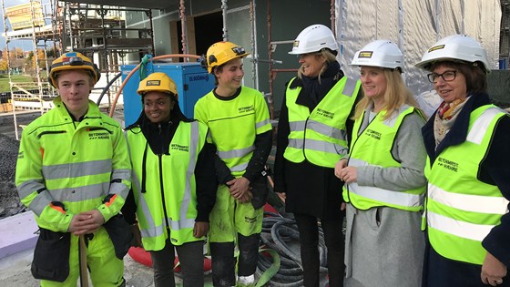 Kari Sandberg i EBA og Sigrun Vågeng i NAV saman med lærlingane og arbeids- og sosialminister Anniken Hauglie. Alle i vernehjelmer og grønt arbeidstøy/vester, stående på byggeplass.