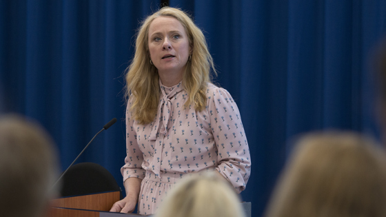 Anniken Hauglie står og snakker for forsamling, blå gardin i bakgrunnen.