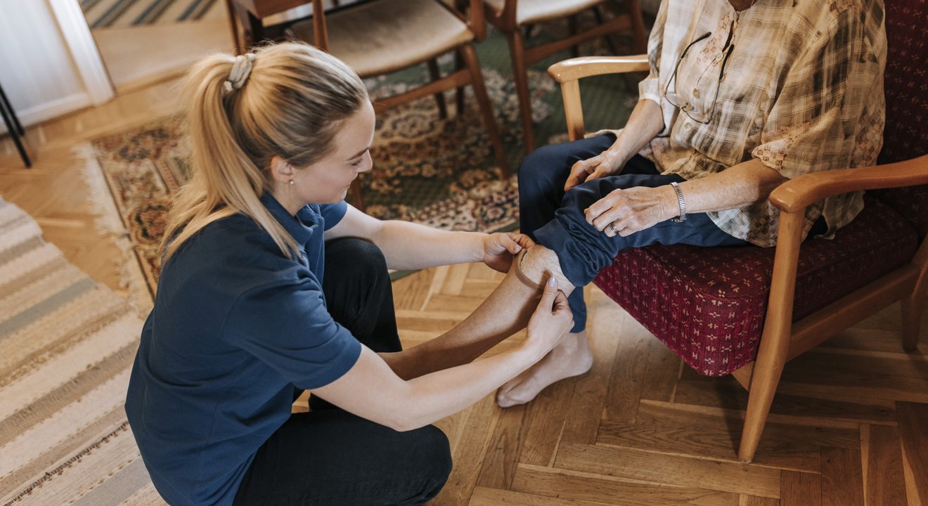 Ei ung jente frå hjemmetenesta som hjelper ei eldre dame med å få på seg strømpene