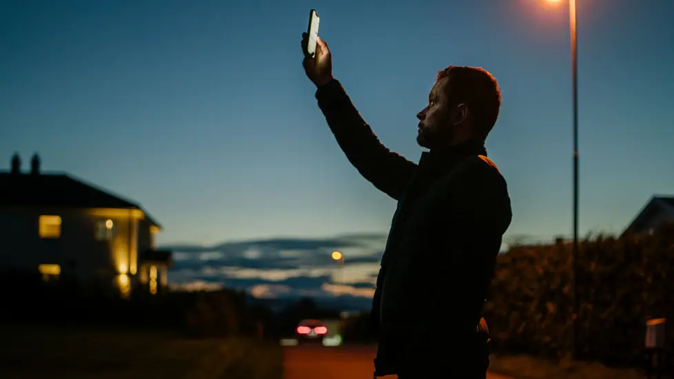 Mann holder mobiltelefonen opp mot himmelen i skumringen.