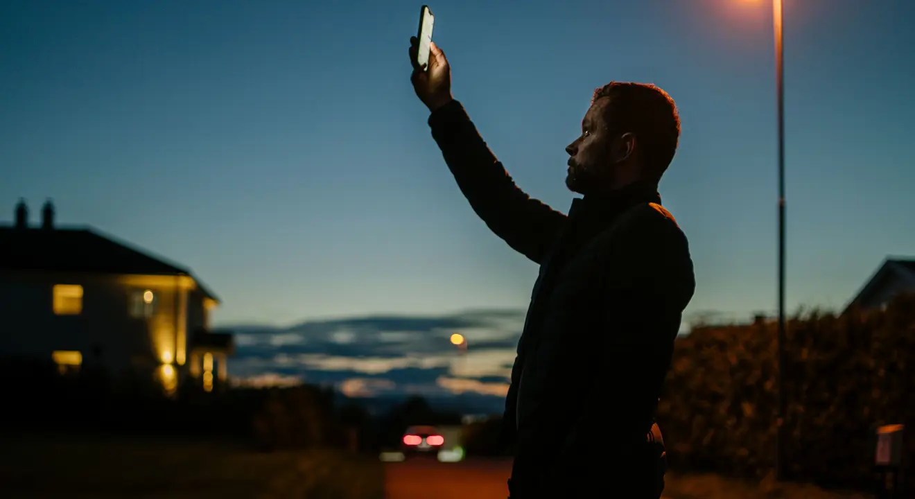 Mann holder mobiltelefonen opp mot himmelen i skumringen.