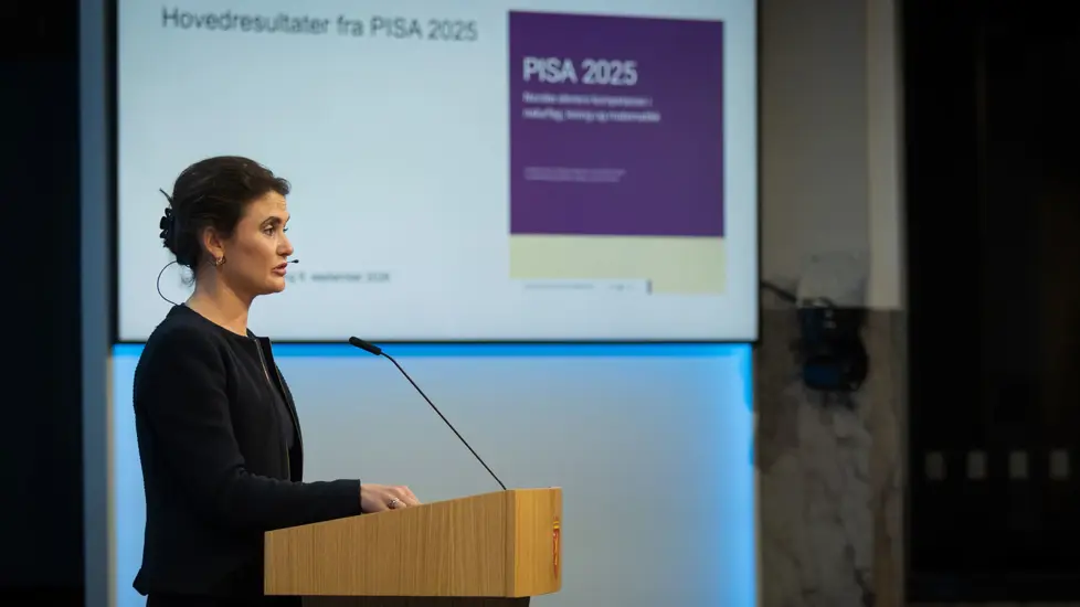 Kari Nessa Nordtun på talerstol. Presentasjon med hovedresultater fra PISA 2025 på skjerm i bakgrunnen.