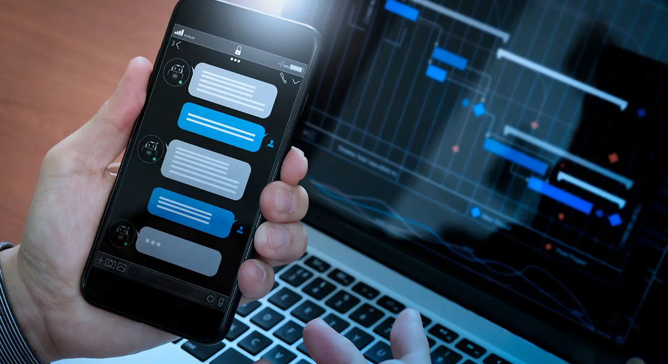 En hånd holder en mobiltelefon med en chattedialog på skjermen - en laptop er i bakgrunnen