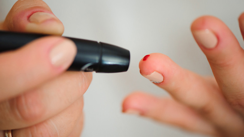 Nærbilde av to hender. En finger har litt blod etter å ha blitt testet med et lite diabetestest-apparat, som man ser på venstre side av bildet.