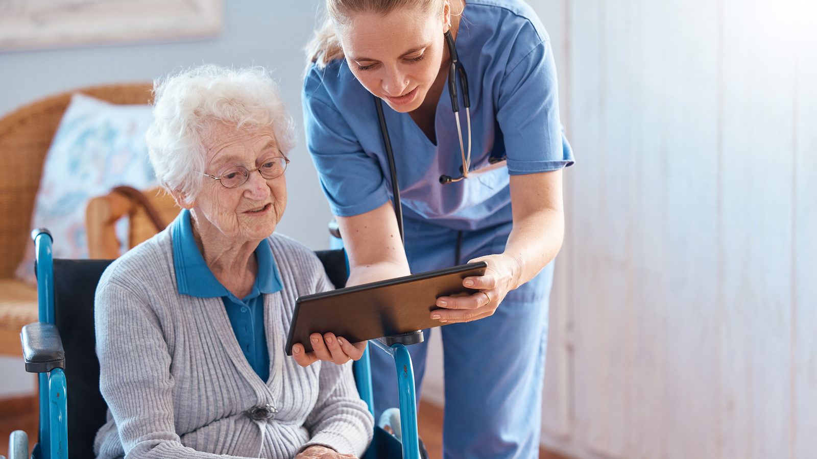 En hjemmepleier viser en eldre person i rullestol et nettbrett