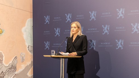 Kvinne med lyst hår og mørk dress står bak et bord med en mikrofon. Hun holder noen ark og står foran en blå vegg med riksløven.