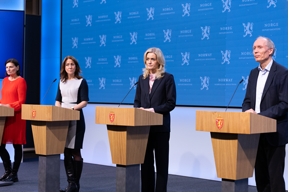 Fire personer bak hver sin talerstol på en scene. Offisiell blå pressevegg med den norske riksløven i bakgrunnen.