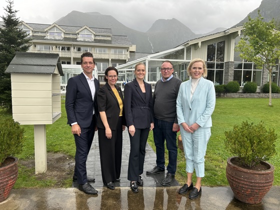 Five people standing next to each other in front of a large wooden house. Misty mountains in the background.
