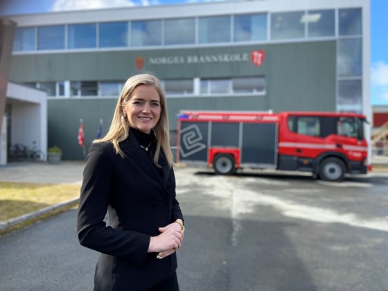 Emilie Enger Mehl står foran en brannbil og lokalene til Norges brannskole.