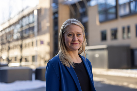 Kvinne med langt blondt hår og blå jakke foran en murbygning. Det er sol og noe snø på bakken.