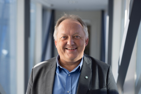 Portrett av smilende mann i grå dress og blå skjorte i lyst kontorlandskap.