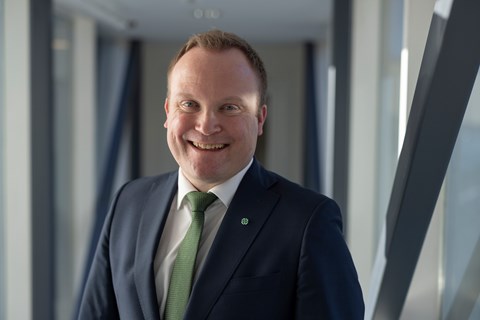 Smilende mann i mørk blå dress og grønt slips som står i lyst kontormiljø.