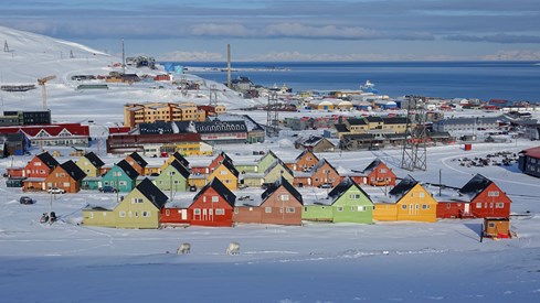 Fargerike hus og diverse bygninger i snølandskap sett skrått ovenfra. Blått hav i bakgrunnen.