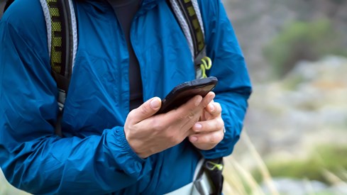 Nærbilde av mann i blå fritidsjakke som ser på en smarttelefon. Han er ute i naturen.