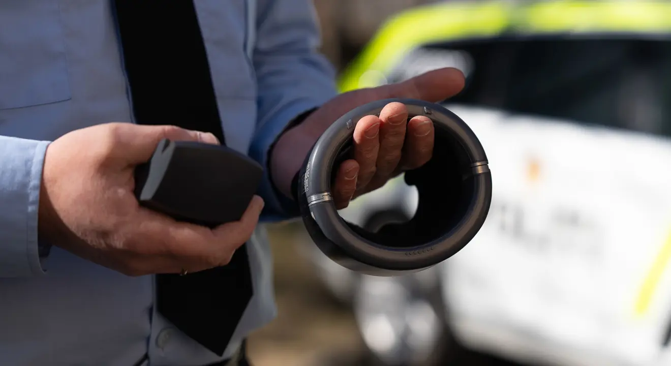 Person i lys blå skjorte og mørkt slips holder fram hendene. I venstre hånd holder han en grå ring og i høyre hånd et grått apparat. I bakgrunnen ser vi en hvit og signalgul bil.