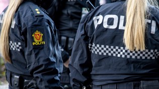 To personer i mørk blå uniform med påskrift "Politi". Begge personene står med ryggen til og har blonde hestehaler nedover ryggen.