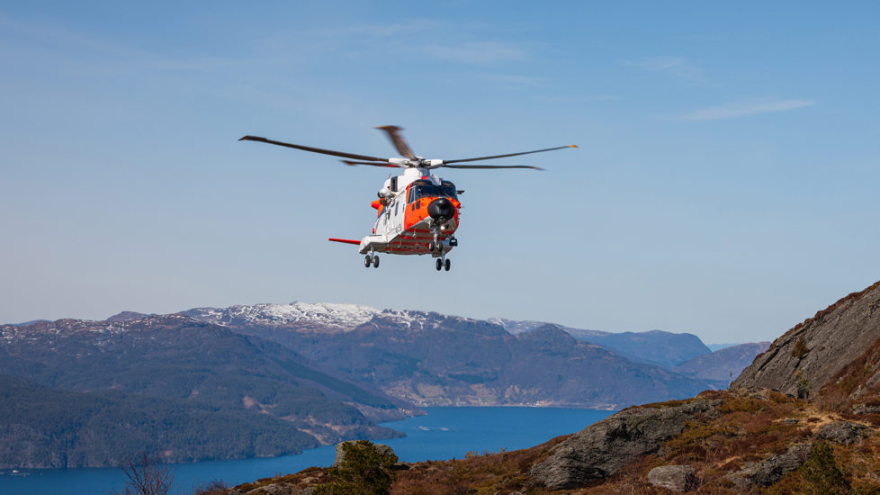 Orange og hvitt helikopter i lufta. Fjell og fjord i bakgrunnen.
