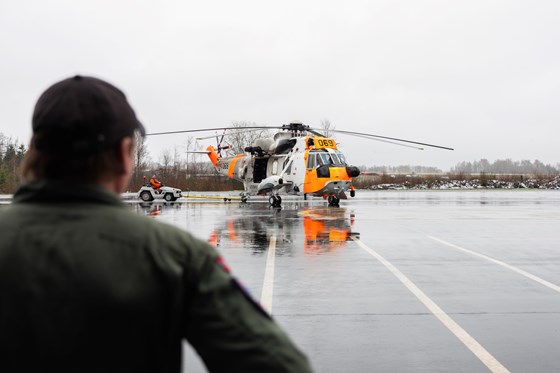 Mann med ryggen til ser på redningshelikopter ute på en landingsplass. Går skyer og litt skog i bakgrunnen.