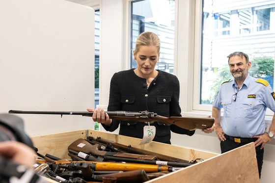 Kvinne i sort drakt holder et gevær foran en kasse med våpen. Politimann i bakgrunnen.