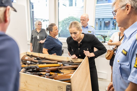 Kvinne i sort drakt holder et gevær foran en kasse med våpen. Flere mennesker, inkludert noen i politiuniform, står rundt