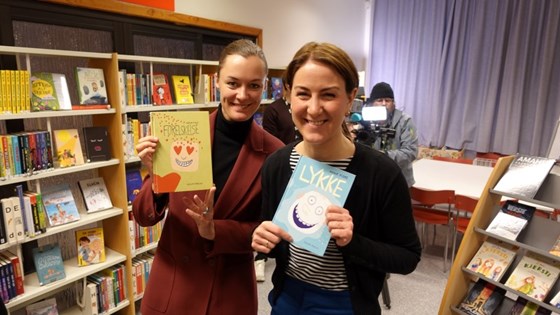 Anette Trettebergstuen og Tonje Brenna holder hver sin bok og smiler til kamera i et skolebibliotek. 