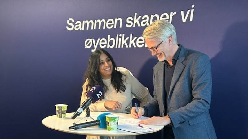 Kultur- og likestillingsminister Lubna Jaffery og sjefsredaktør i TV 2 Olav T. Sandnes signerer den nye avtalen, stående ved et høyt kafébord med blå vegg bak hvor det står "Sammen skaper vi øyeblikkene".