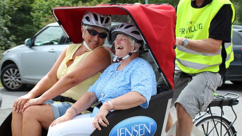 Frivillig som sykler en sykkelvogn med to voksne passasjerer.