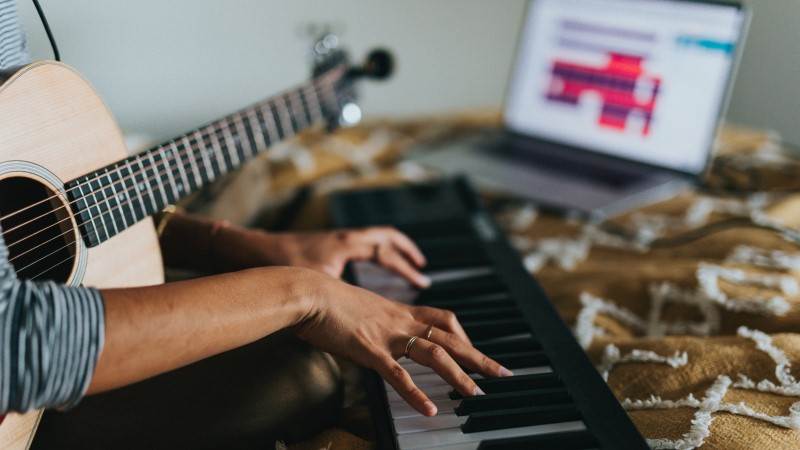 En person spiller gitar og keyboard med en PC i bakgrunnen