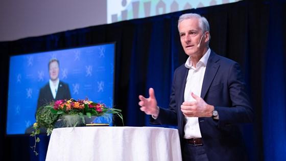 Statsminister Jonas Gahr Støre taler på scenen bak et rundt bort med duk. Kommunalminister Gjelsvik er med digitalt og vises på veggen bak ham.