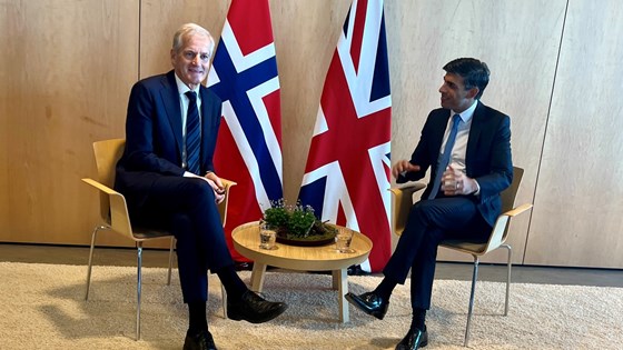 Statsminister Støre og Storbritannias statsminister Sunak sitter på hver sin side av et rundt bord med nasjonenes flagg som bakgrunn.