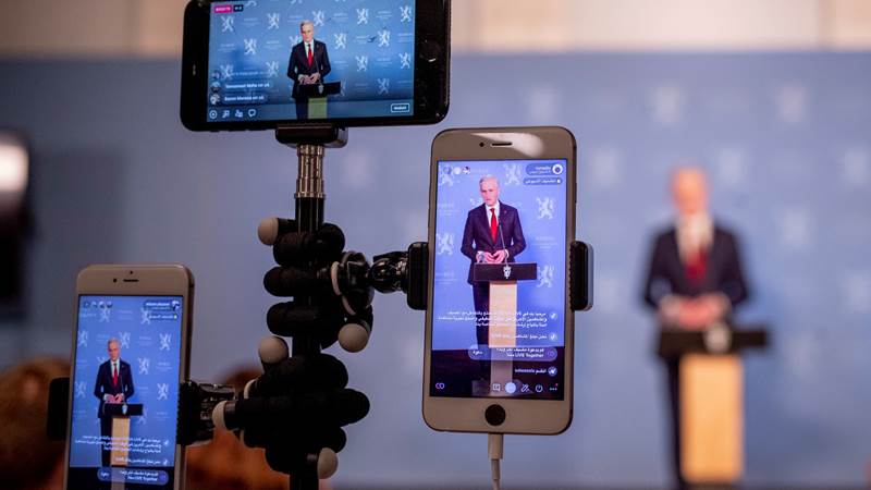 Tre mobiltelefoner i et stativ - alle peker mot en blåvegg med statsminister Jonas Gahr Støre i forgrunnen.