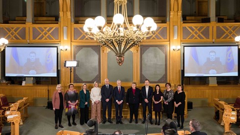 Gruppebilde av partiledere og to parlamentariske ledere i Lagtingsssalen. President Zelenskyj på skjerm i bakgrunnen.