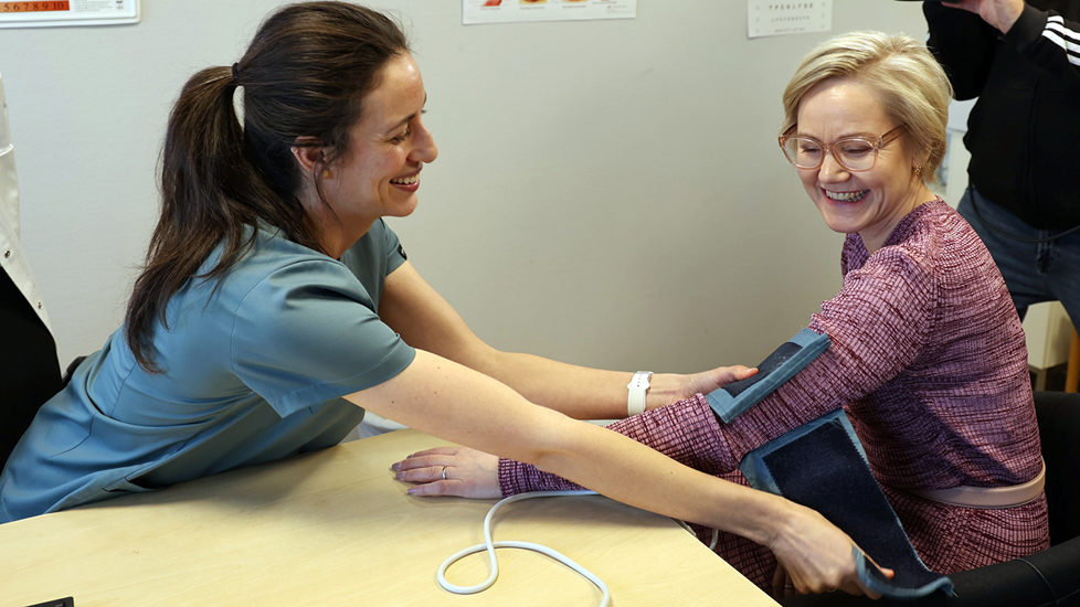 En fastlege utfører en måling på helse- og omsorgsminister Ingvild Kjerkol mens begge smiler
