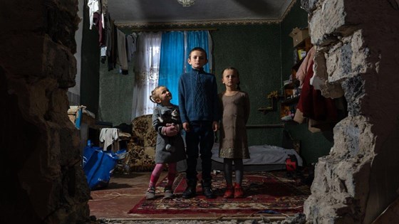 Tre søsken i et rom som har skader etter et angrep i Ukraina. Hull i veggen.