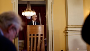 Statsministeren står bak en talerstol. Lyskrone i bakgrunnen.