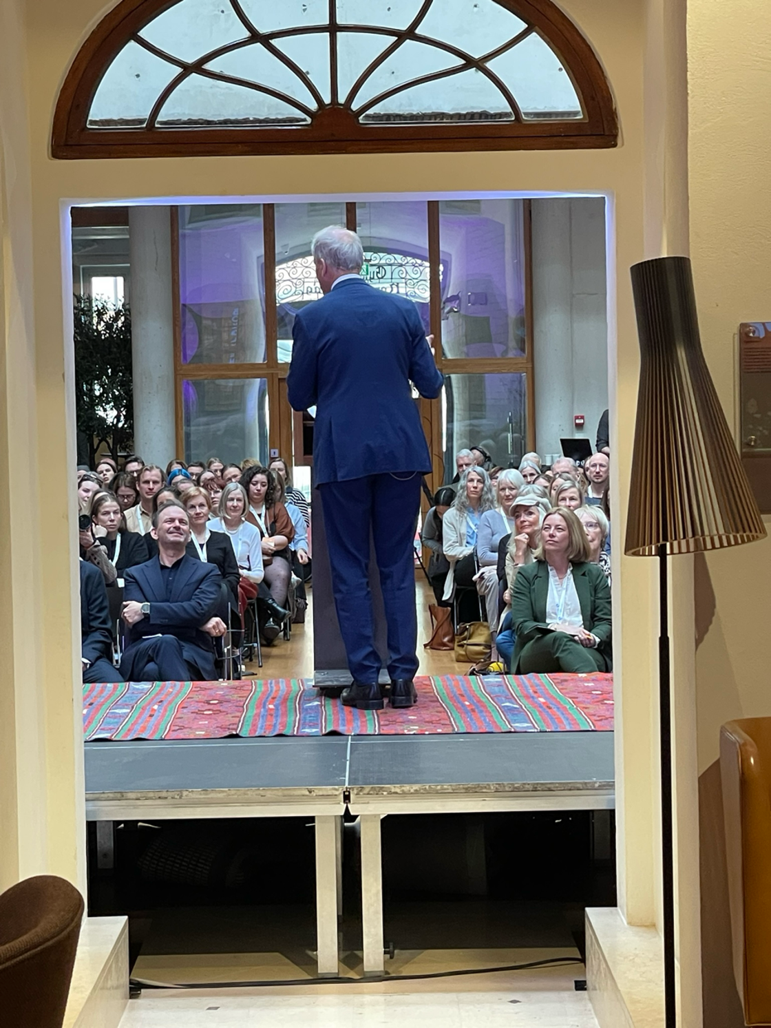 Statsministeren står på en scene med ryggen til. Publikum sitter på stoler og ser mot scenen.