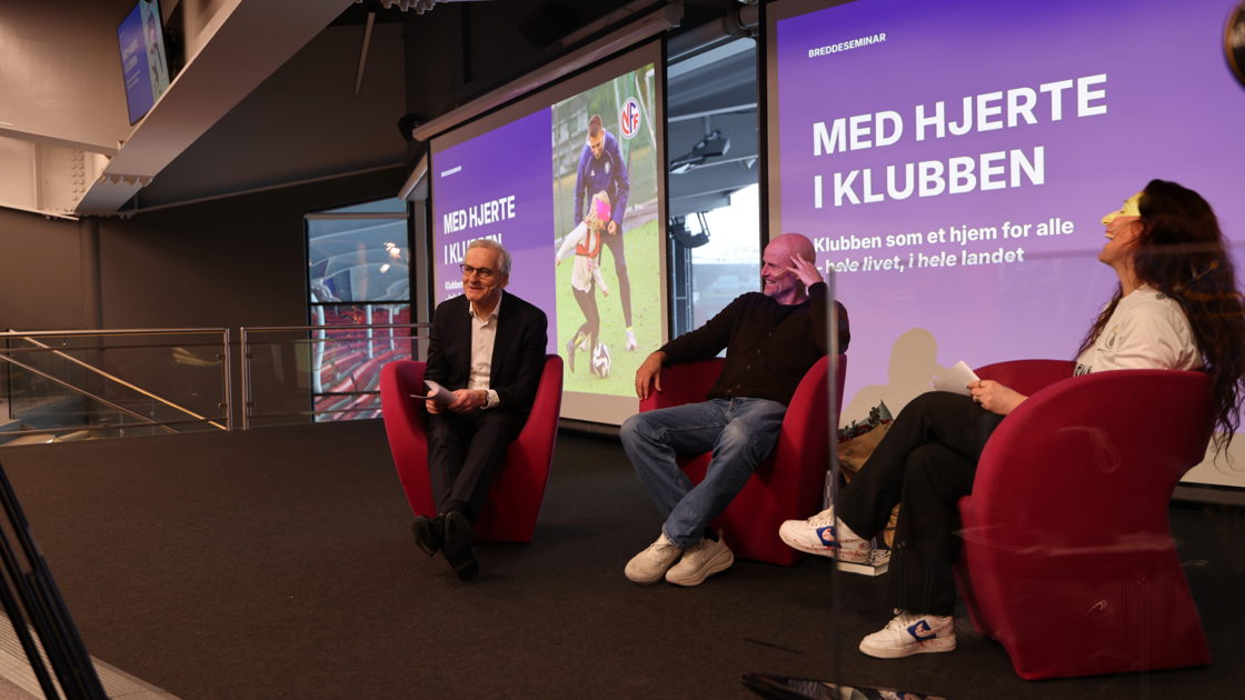 De tre sitter i røde stoler på en scene foran to digitale skjermer med teksten "Med hjerte i klubben"