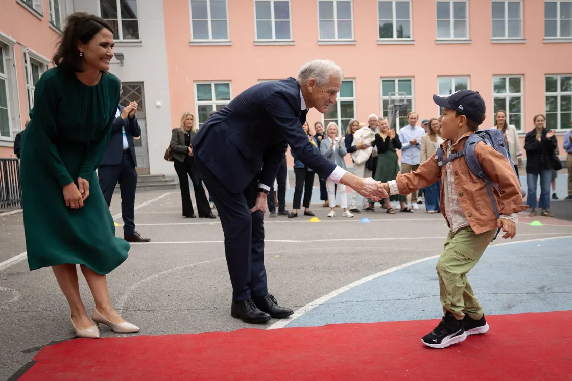 Statsministeren tar en førsteklassing i hånda. Førsteklassingen går over rød løper på vei inn til klasserommet.