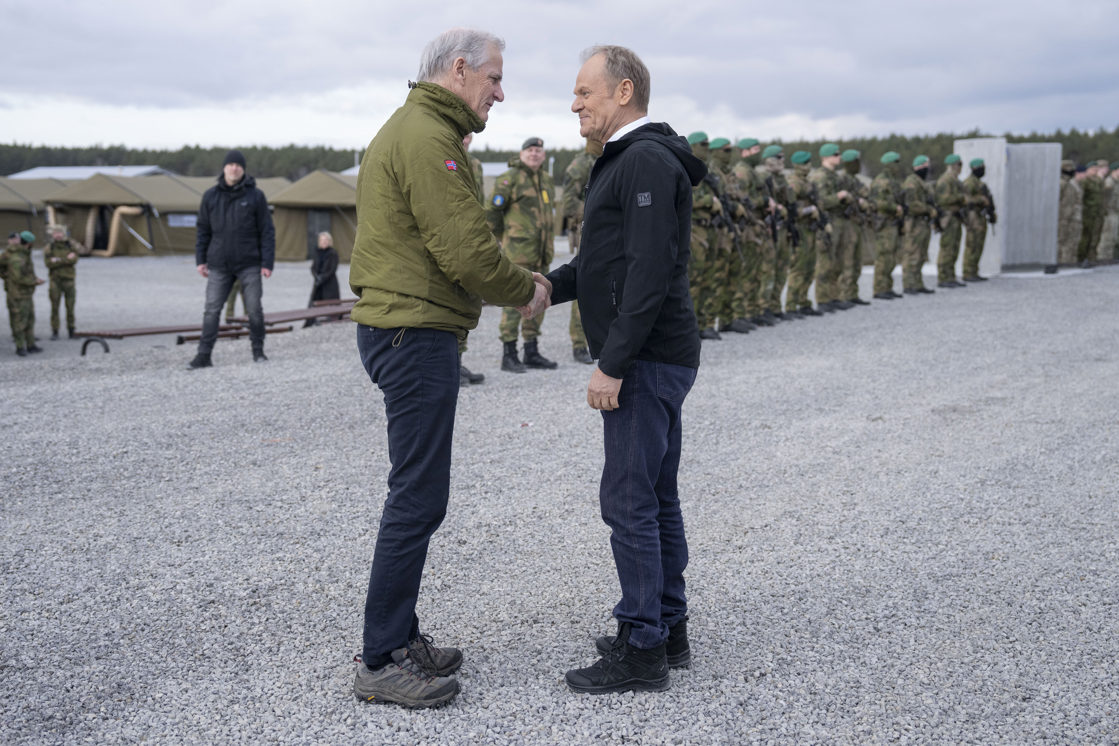 Jonas Gahr Støre hilser på Donald Tusk i en militær treningsbase. Soldater oppstilt i bakgrunnen.