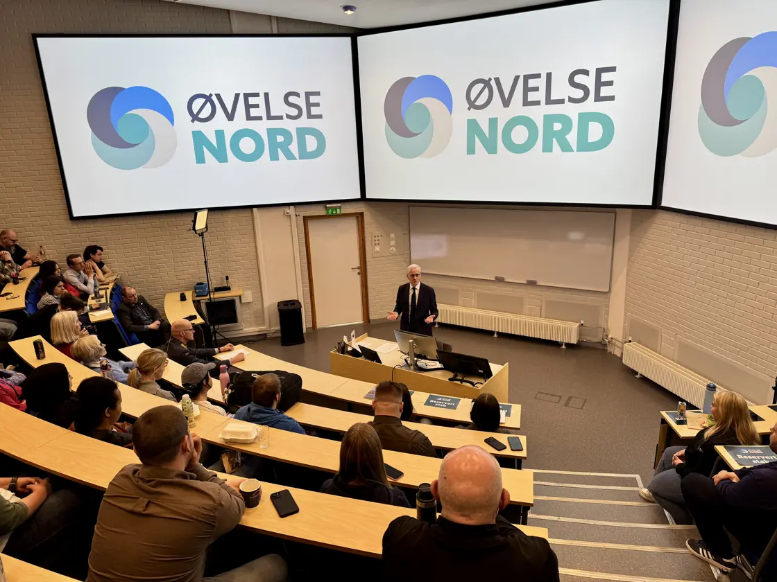 Statsministeren står i et auditorium. Publikum sitter på stoler oppover i amfiet.