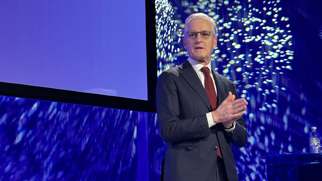 Statsministeren står på scenen. Holder håndflatene mot hverandre. Blå dress med mørkerødt slips og hvit skjorte.