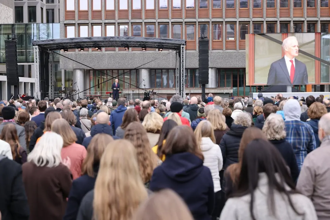 Statsministeren står på scenen foran høyblokka. Publikum står foran scenen.