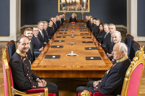 Statsrådsmøte på Slottet. Regjeringen sitter samlet rundt et stort møtebord, men Kongen og Kronprinsen nærmest kamera.